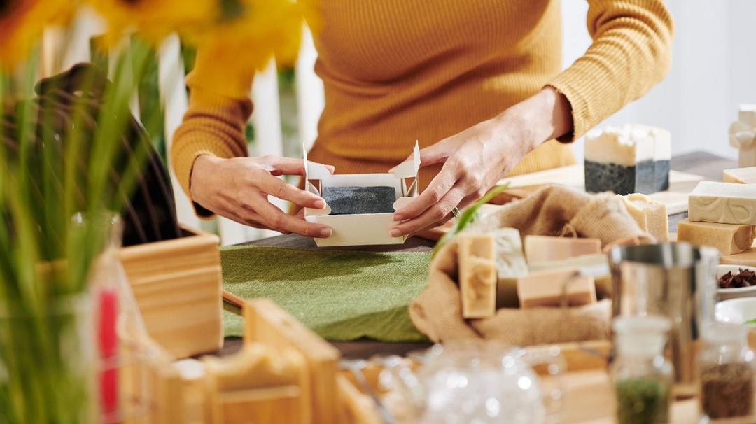 A person in a yellow sweater packaging a handmade soap bar at a craft table surrounded by various natural ingredients and materials.