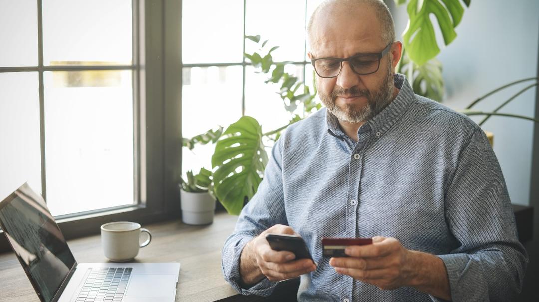 A balding middle-aged man with glasses, using a smartphone and holding a credit card, seated at a desk with a laptop and coffee cup nearby.